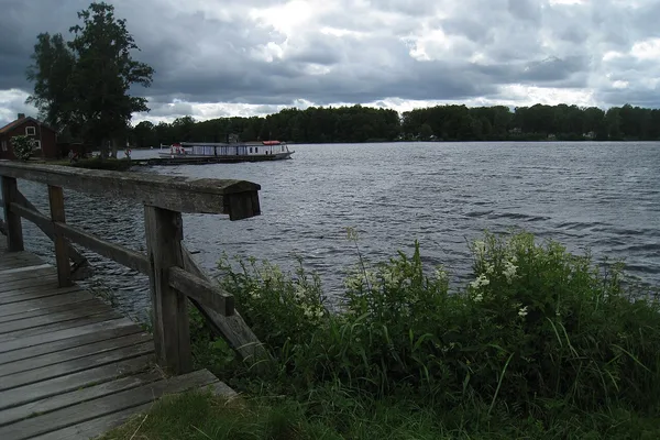 Träbrygga vid sjö med ångbåt, grönt gräs i förgrunden