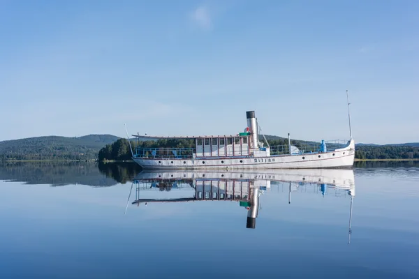 Ångbåten Siljan speglar sig i stilla sjövatten med berg bakom