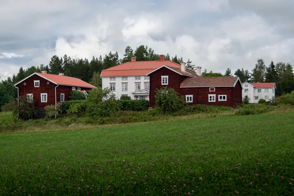 Hälsingegård med röda timmerbyggnader och vit herrgård på äng